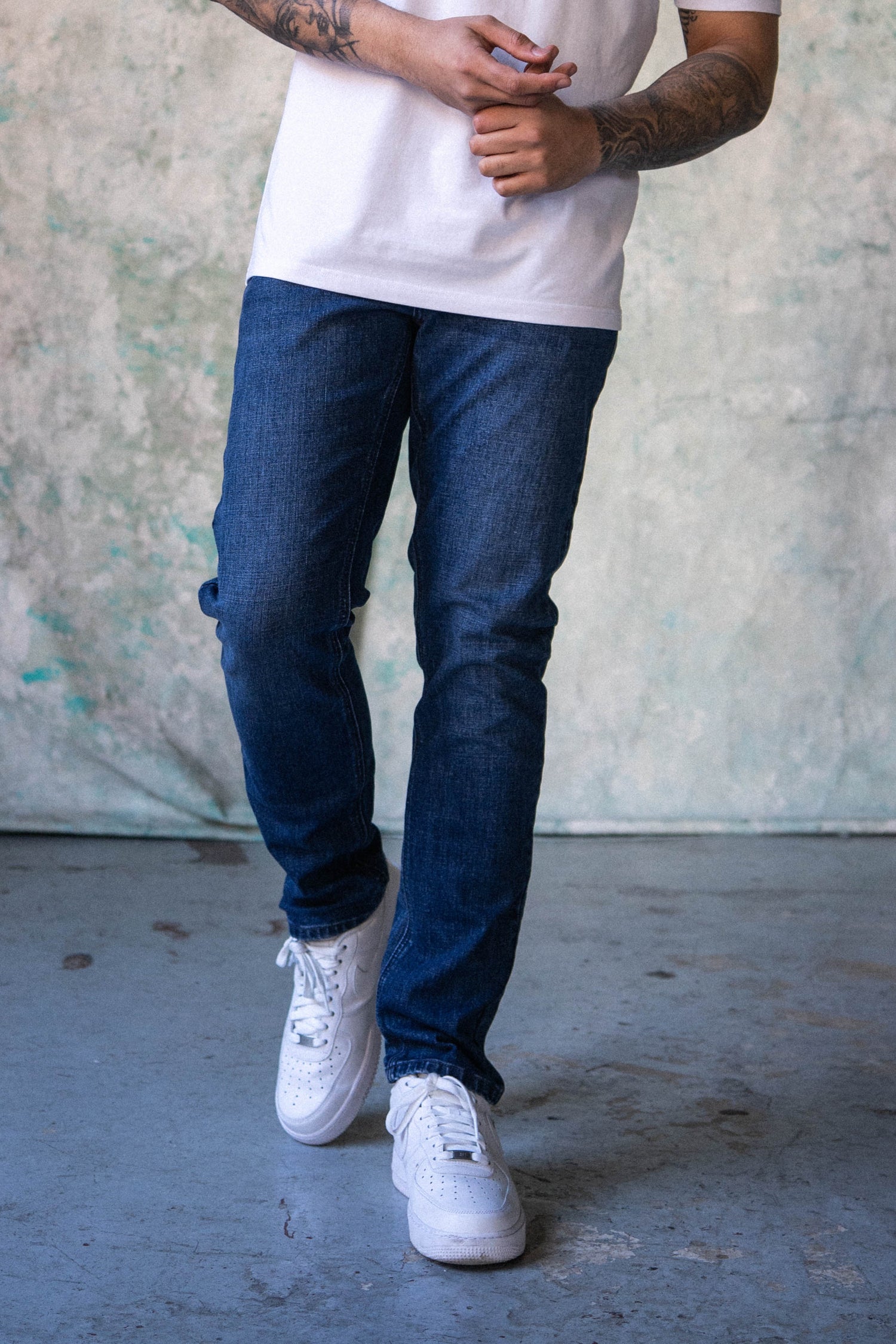 Man wearing a white t-shirt and blue jeans standing against a textured wall.